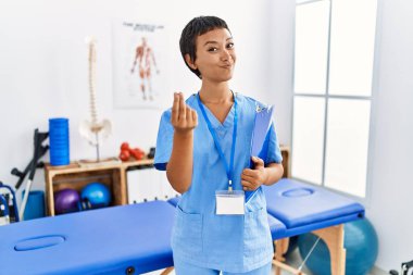 Kısa saçlı, İspanyol bir kadın. Ağrı giderme kliniğinde çalışıyor. Elleriyle para jesti yapıyor. 