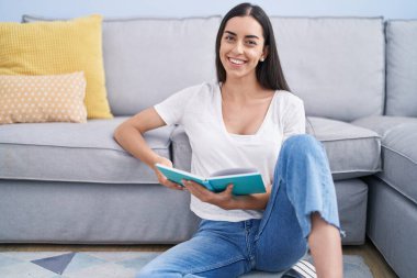 Young hispanic woman reading book sitting on floor at home