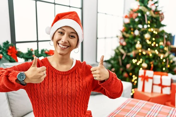 Kısa saçlı, kısa saçlı, Noel şapkalı genç bir kadın kanepeye oturmuş, başarı işareti yapıyor, elleriyle olumlu jest yapıyor, gülümsüyor ve mutlu. Neşeli ifade ve kazanan jest. 