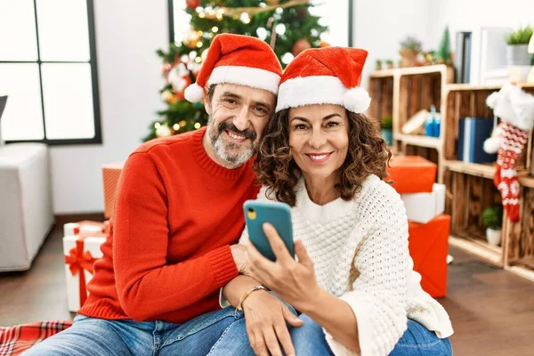 中世のヒスパニック系のカップルの幸せなクリスマスの帽子を身に着けて笑顔 自宅でスマホを使って床に座る — ストック写真