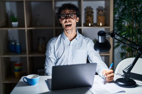 Hispanic man working at the office at night angry and mad screaming frustrated and furious, shouting with anger. rage and aggressive concept.