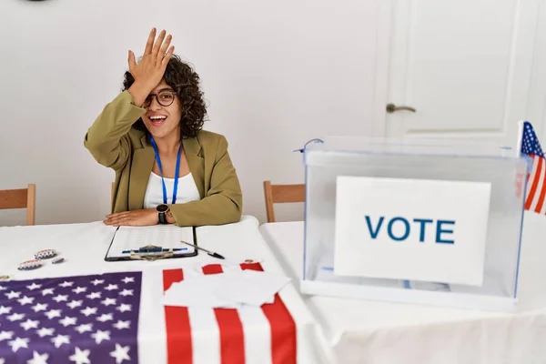 Young hispanic woman at political election sitting by ballot surprised with hand on head for mistake, remember error. forgot, bad memory concept.