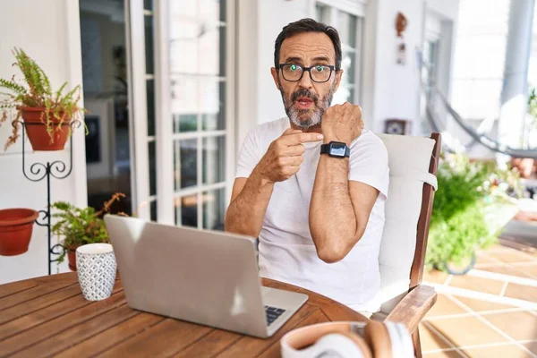 Middle age man using computer laptop at home in hurry pointing to watch time, impatience, looking at the camera with relaxed expression