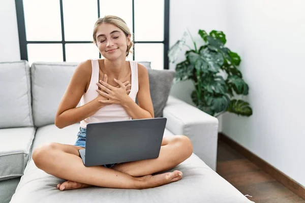 年轻的金发女人在家里用笔记本电脑坐在沙发上 双手放在胸前笑着 闭着眼睛 脸上挂着感激的手势 健康概念 — 图库照片