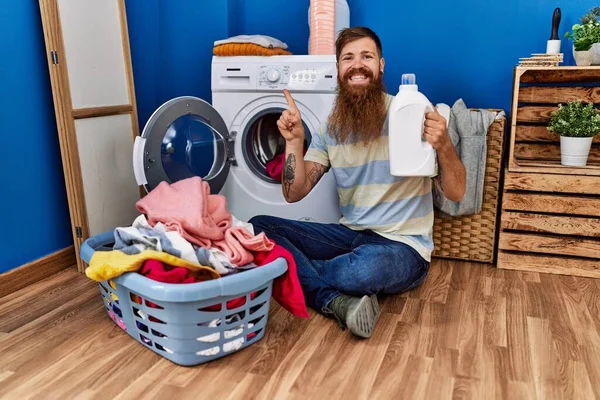 Redhead man with long beard doing laundry holding detergent bottle smiling happy pointing with hand and finger to the side