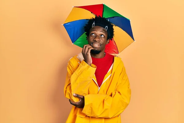 若いアフリカ系アメリカ人の男性は 顎の質問について考える上で手で黄色のレインコートを着て 集中的な表現 笑顔で思いやりのある顔 疑わしい概念 — ストック写真