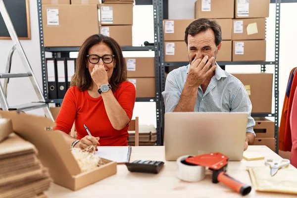 Middle age couple working at small business ecommerce smelling something stinky and disgusting, intolerable smell, holding breath with fingers on nose. bad smell