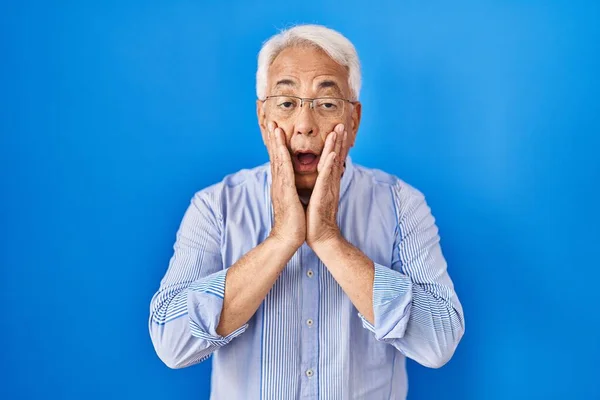 Hispanic senior man wearing glasses afraid and shocked, surprise and amazed expression with hands on face