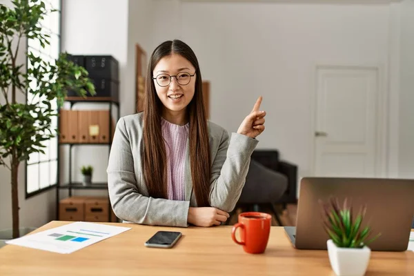 Young chinese business worker wearing business style sitting on desk at office smiling happy pointing with hand and finger to the side