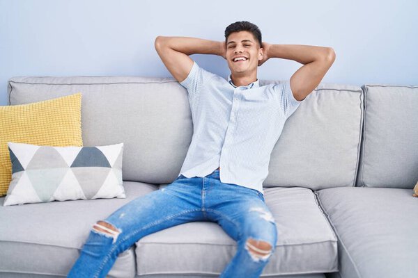 Young hispanic man relaxed with hands on head sitting on sofa at home