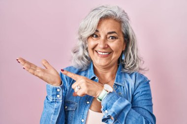 Pembe arka planda duran gri saçlı orta yaşlı kadın kameraya hayret ediyor ve gülümsüyor.. 