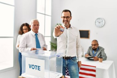 Orta yaşlı Amerikalı seçmen gülümseyerek rozetini oy merkezinde tutuyor..