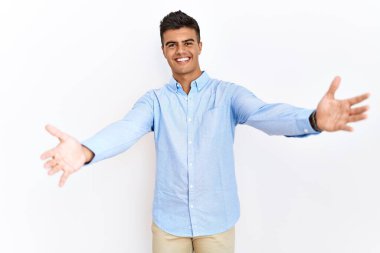 İş gömleği giyen genç İspanyol bir adam izole edilmiş arka planda durup kameraya bakarak sarılmak için kollarını açmış gülümsüyor. Mutluluğu kucaklayan neşeli bir ifade.. 