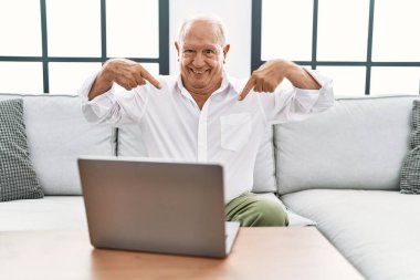 Evde dizüstü bilgisayar kullanan kıdemli bir adam kanepede oturuyor. Kendine güveni tam görünüyor. Kendini gururlu ve mutlu bir şekilde gösteriyor.. 