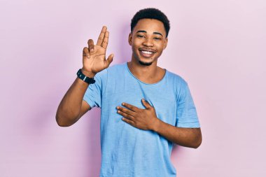 Genç, Afro-Amerikan bir adam, günlük mavi tişört giyiyor, gülümsüyor, elleri göğsünde ve parmakları havada küfür ediyor, sadakat yemini ediyor. 