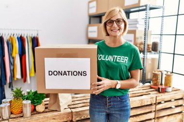 Middle age blonde woman wearing volunteer t shirt holding donations box looking positive and happy standing and smiling with a confident smile showing teeth 