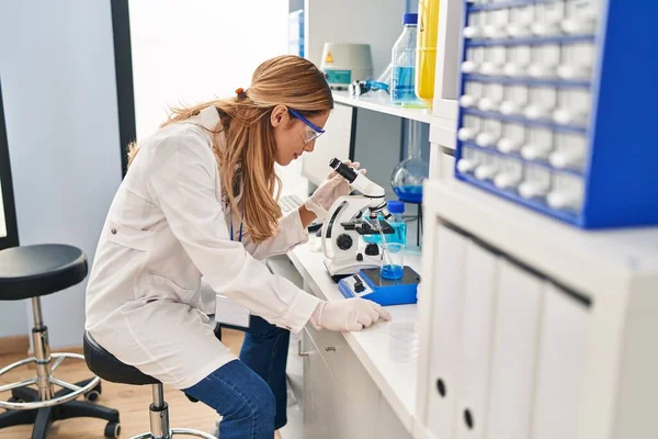 实验室显微镜下身穿科学家制服的金发年轻女子 — 图库照片