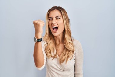 İzole edilmiş arka planda duran genç sarışın kadın kızgın ve kızgın yumruğunu kaldırıyor ve öfkeyle bağırıyor. öfke ve agresif konsept. 
