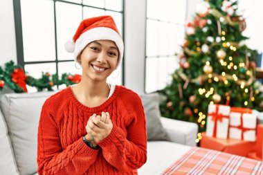 Kısa saçlı, ispanyol bir kadın, Noel şapkası takıyor, kanepede oturuyor, ellerini kavuşturup gülümsüyor ve gülümsüyor. Başarı ve iyimserlik 