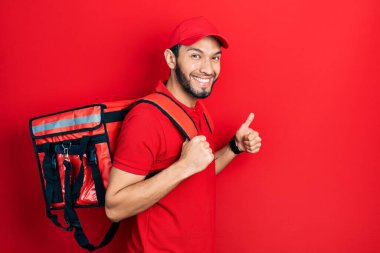 Hispanic man with beard wearing delivery package backpack smiling happy and positive, thumb up doing excellent and approval sign 
