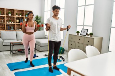 Genç latin çift mutlu bir şekilde gülümsüyor. Evde elastik bant ve dambıl kullanıyorlar..