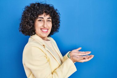 Young brunette woman with curly hair standing over blue background pointing aside with hands open palms showing copy space, presenting advertisement smiling excited happy 