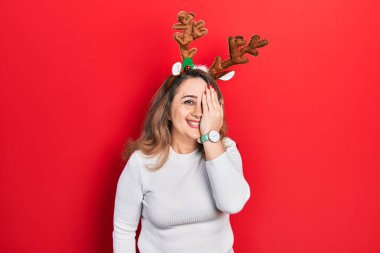 Middle age caucasian woman wearing cute christmas reindeer horns covering one eye with hand, confident smile on face and surprise emotion. 
