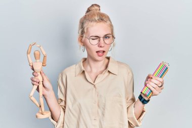 Beautiful caucasian woman with blond hair holding small wooden manikin pencils winking looking at the camera with sexy expression, cheerful and happy face. 