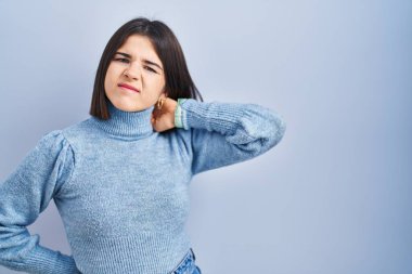 Mavi arka planda dikilen genç İspanyol kadın boyun ağrısı çekiyor, eliyle boynuna dokunuyor, kas ağrısı çekiyor. 