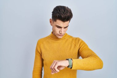 Mavi arka planda duran genç İspanyol adam kol saatindeki saati kontrol ediyor, rahat ve kendinden emin. 