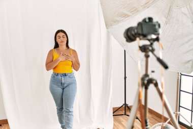 Fotoğraf stüdyosunda model olarak poz veren genç, güzel İspanyol bir kadın elleri göğsünde, gözleri kapalı ve yüzünde minnet dolu bir jest ile gülümsüyor. sağlık kavramı. 