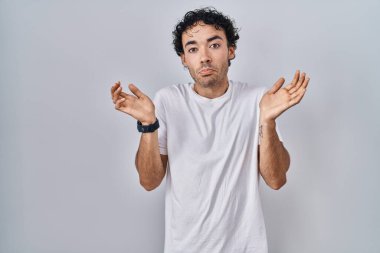 Hispanic man standing over isolated background clueless and confused expression with arms and hands raised. doubt concept. 