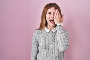 Beautiful woman standing over pink background covering one eye with hand, confident smile on face and surprise emotion. 