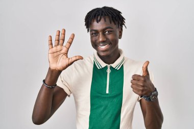 Afrikalı bir adam izole edilmiş arka planda duruyor. Altıncı parmaklarını gösterip işaret ediyor. Kendinden emin ve mutlu bir şekilde gülümsüyor.. 