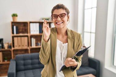 Young woman working at consultation office gesturing finger crossed smiling with hope and eyes closed. luck and superstitious concept. 