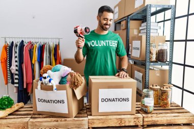 Young hispanic man wearing volunteer uniform packing donations box at charity center