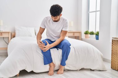 Genç Arap adam diz sakatlığından muzdarip. Yatak odasında oturuyor.