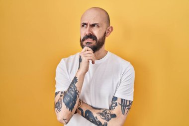 Young hispanic man with tattoos standing over yellow background thinking worried about a question, concerned and nervous with hand on chin 