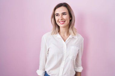 Young beautiful woman standing over pink background smiling looking to the side and staring away thinking. 