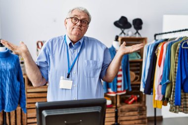 Senior man with grey hair working as manager at retail boutique clueless and confused expression with arms and hands raised. doubt concept. 