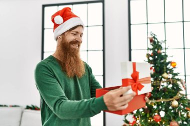 Genç kızıl saçlı boks hediyesini çıkaran adam Noel ağacının yanında duruyor.