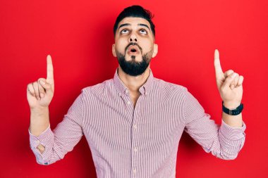 Handsome man with beard wearing casual red shirt amazed and surprised looking up and pointing with fingers and raised arms. 