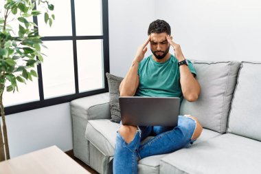 Sakallı genç yakışıklı adam evdeki koltukta oturan bilgisayarlı dizüstü bilgisayarını kullanıyor ve stres yüzünden başı ağrıyor. migren ağrısı. 