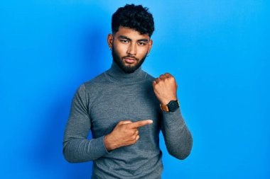Arab man with beard wearing turtleneck sweater in hurry pointing to watch time, impatience, looking at the camera with relaxed expression 