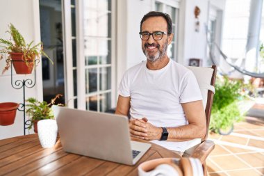 Orta yaşlı bir adam evde bilgisayar kullanıyor. Elleri birarada ve gülümsüyor. Rahatlamış ve neşeli. Başarı ve iyimserlik 