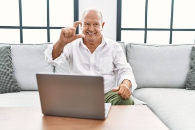 Evde dizüstü bilgisayar kullanan, koltukta oturan, gülümseyen ve kendine güvenen, el hareketi yapan, parmaklarıyla işaret yapan, kameraya bakan yaşlı bir adam. ölçüm kavramı. 