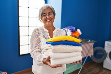 Çamaşırhanede katlanmış giysileri tutan orta yaşlı bir kadın gülümsüyor.