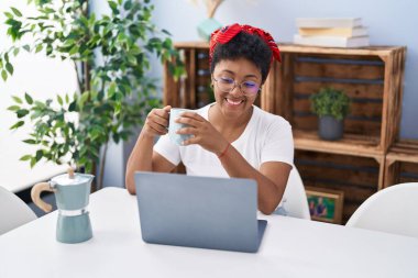 Laptop kullanan ve kahve içen Afro-Amerikalı bir kadın evde oturuyor.