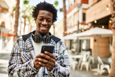 Afro saçlı, kulaklık takan yakışıklı, açık havada mutlu bir şekilde gülümseyen akıllı telefon kullanan bir zenci.