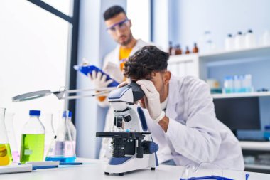 Mikroskop kullanan iki bilim adamı laboratuarda çalışan panoya yazı yazıyorlar.